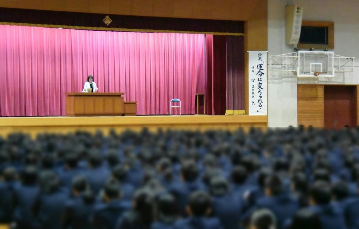 母校・高松商業高等学校の体育館の舞台で講演する宙SORA。垂れ幕に演題「運命は変えられる！」